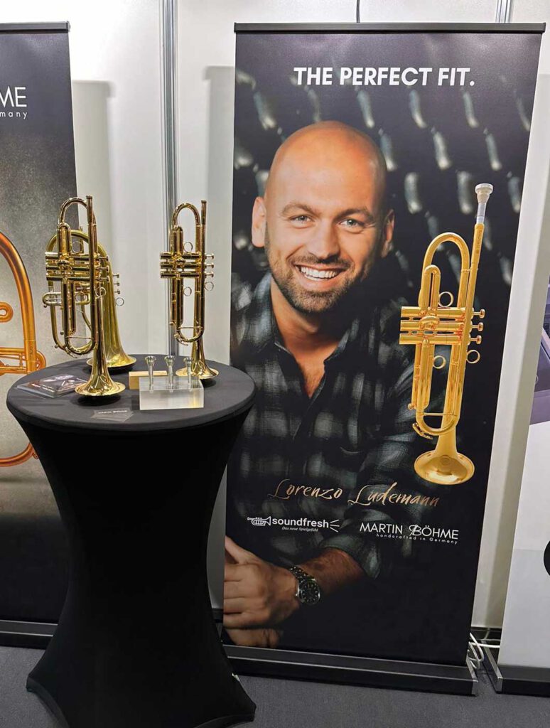 Messestand rangezoomt, im Bild ist Roll-up mit Titel the perfect fit. und Lorenzo Ludemann mit Trompete abgebildet. Davor steht ein schwarzer runder Stehtisch mit Trompeten und Mundstücken drauf.